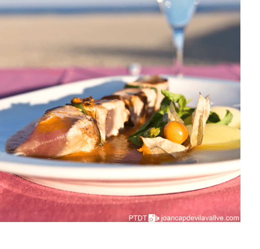 Fotografia on es veu una taula d'un restaurant amb un plat de menjar. De fons s'intueix la platja.